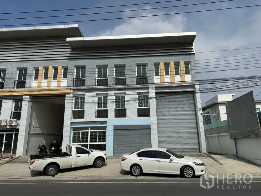 Factory for rent in Bang Sao Thong, Samut Prakan - Three-storey factory-warehouse facade with roller shutter doors and roadside parking in Bang Sao Thong.