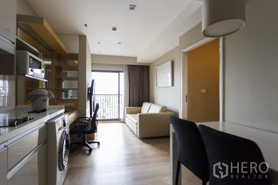 Condominium for rent in Khlong Toei, Bangkok - Bright living area with sofa, work desk, balcony doors and an open kitchenette with washer.