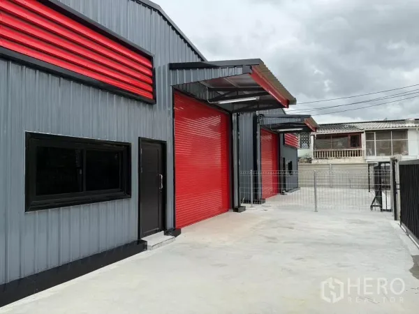 Warehouse for rent in Lat Phrao, Bangkok - Side view of steel-clad warehouse showing pedestrian door, windows, and multiple red roller shutters.