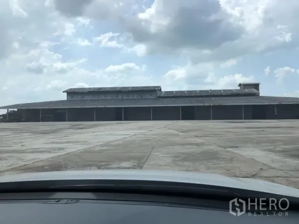 货栈 出租在Chaloem Phra Kiat, Saraburi - 沙拉武里大型金属仓库，多个卷闸装卸门与宽阔混凝土堆场，天空多云。