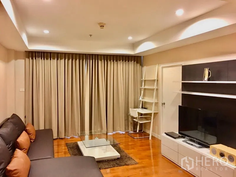 Condominium for rent in Khlong Toei, Bangkok - Living area with sectional sofa, TV cabinet, study desk and floor-to-ceiling curtains.