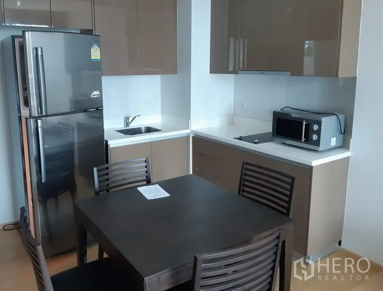 公寓 出租在Khlong Toei, Bangkok - 曼谷空堤区公寓内的L形厨房，亮面橱柜、双门冰箱、微波炉与四人餐桌。