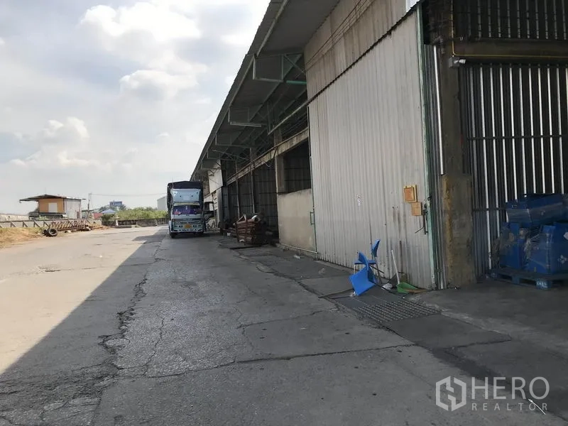 Warehouse for rent in Mueang Pathum Thani, Pathum Thani - Exterior loading area beside the warehouse with truck access and tall sliding metal doors.