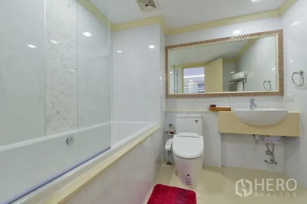 Apartment for rent in Khlong Toei, Bangkok - Modern white bathroom with large mirror, bathtub with glass screen, and toilet.