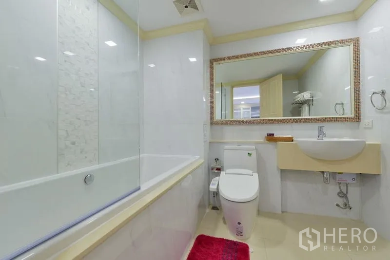 Apartment for rent in Khlong Toei, Bangkok - Modern white bathroom with large mirror, bathtub with glass screen, and toilet.