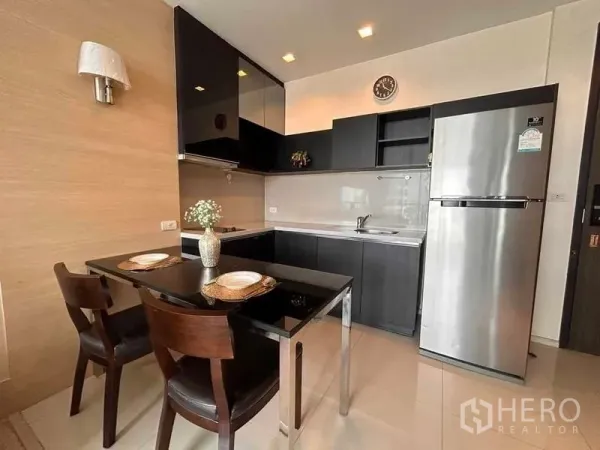 Condominium for rent or sale in Khlong Toei, Bangkok - L-shaped kitchen with sleek black cabinetry, dining table and stainless-steel refrigerator.