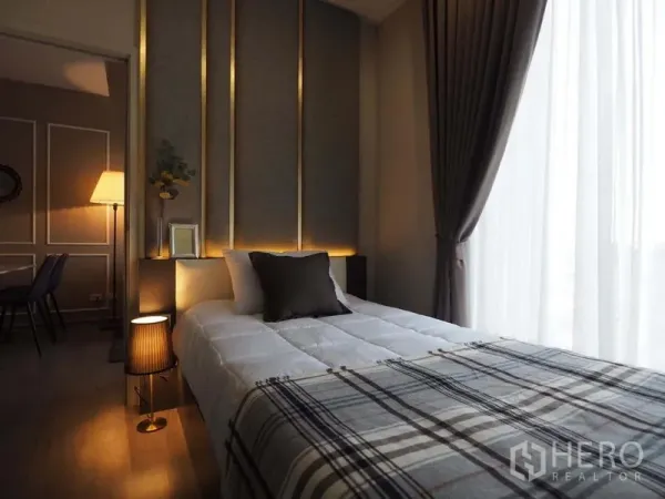 Condominium for rent in Khlong San, Bangkok - Bedroom with padded headboard, warm accent lighting, and floor-to-ceiling window with sheer curtains.