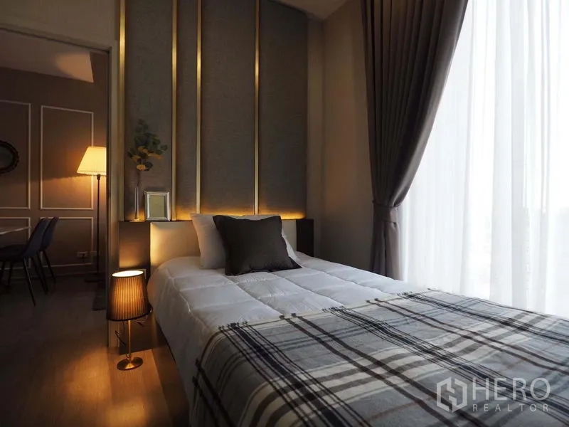 Condominium for rent in Khlong San, Bangkok - Bedroom with padded headboard, warm accent lighting, and floor-to-ceiling window with sheer curtains.