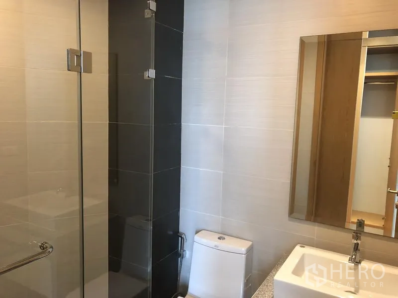 Condominium for rent in Khlong Toei, Bangkok - Contemporary bathroom with glass shower, vanity, mirror and toilet.