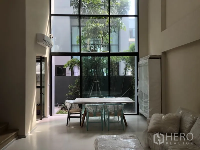 Townhouse for rent in Suan Luang, Bangkok - Dining area with double-height ceiling and floor-to-ceiling glass opening to a small garden.
