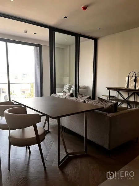 Condominium for sale in Watthana, Bangkok - Open‑plan space with dining set, glass partition to a multipurpose room, balcony access and herringbone wooden flooring.