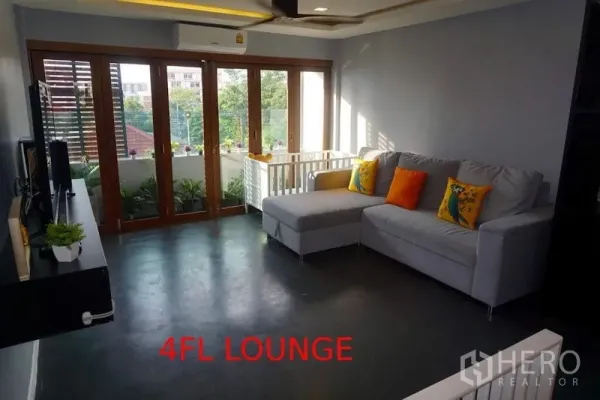 Shophouse for sale in Phra Khanong, Bangkok - Bright lounge with an L-shaped sofa, TV wall and floor-to-ceiling glass doors opening to a balcony.