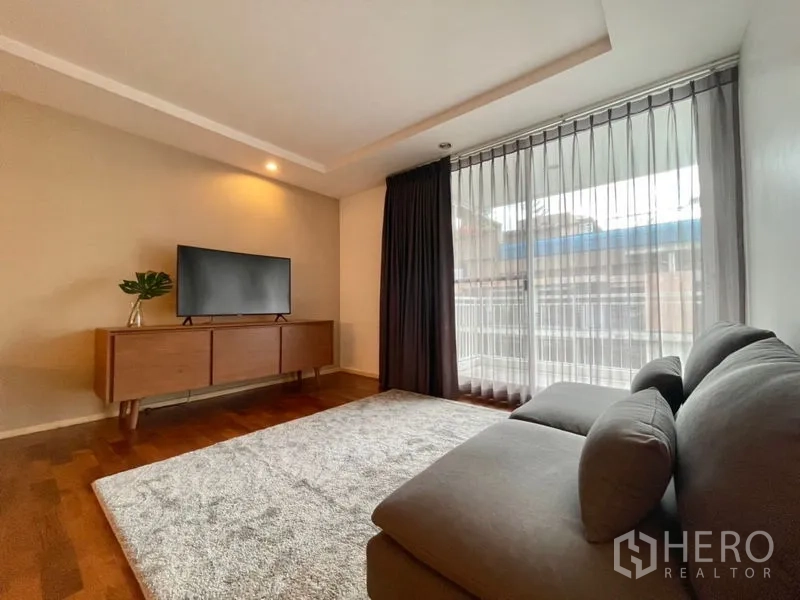Condominium for rent or sale in Khlong Toei, Bangkok - Living area with sectional sofa, TV console, and floor-to-ceiling windows with curtains.