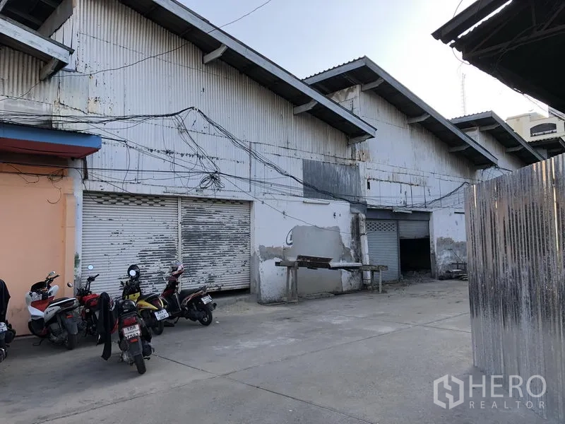 零售空间 出租在Khlong Toei, Bangkok - 波纹金属外立面，多个卷闸门与装卸前院，可见停放的摩托车。