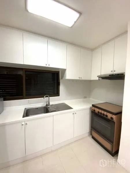 公寓 出租在Watthana, Bangkok - 紧凑白色厨房，上下橱柜齐全，窗下不锈钢水槽，配烤箱与油烟机。