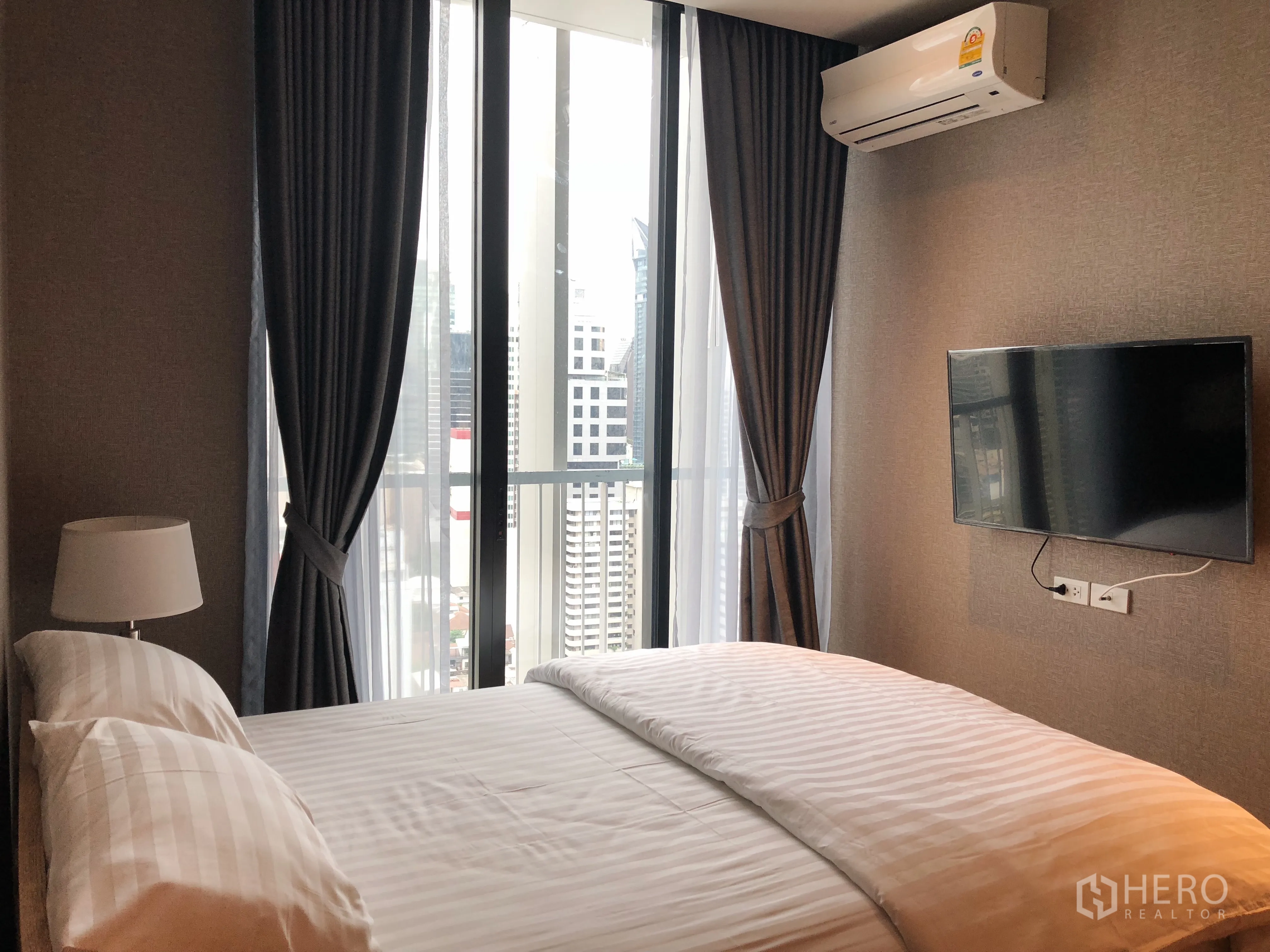 Condominium for rent in Khlong Toei, Bangkok - Cozy bedroom with floor-to-ceiling window, wall-mounted TV and city skyline view.