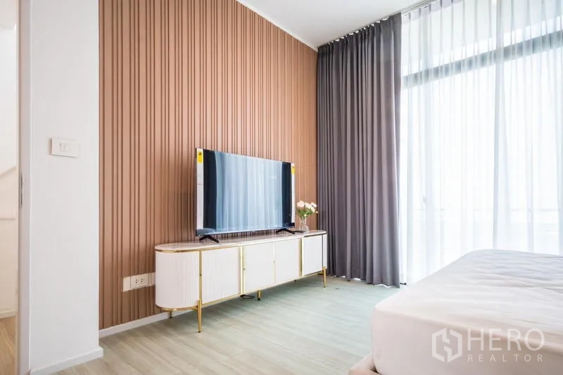 Townhouse for rent or sale in Suan Luang, Bangkok - Feature wall with TV console and full-height curtains beside the bed.