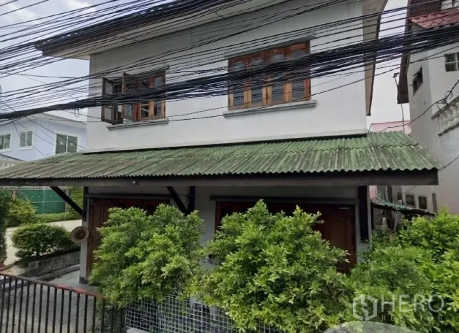 Detached House for sale in Phra Khanong, Bangkok - Two-storey detached house in Phra Khanong with green awning roof, timber windows, gated front garden, and overhead utility lines.