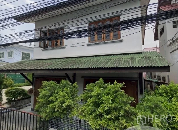 Detached House for sale in Phra Khanong, Bangkok - Two-storey detached house in Phra Khanong with green awning roof, timber windows, gated front garden, and overhead utility lines.
