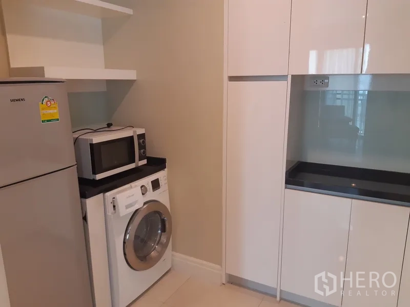 Condominium for rent in Khlong Toei, Bangkok - Modern kitchen with glossy white cabinets, black countertops, fridge, microwave, and front-load washing machine.
