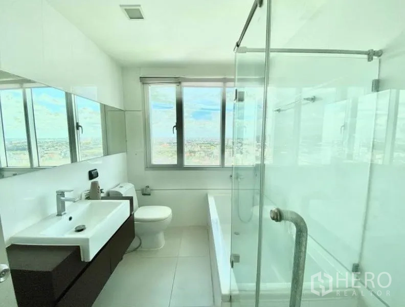 Condominium for rent in Huai Khwang, Bangkok - Bright bathroom with glass shower, bathtub, vanity and wide windows.