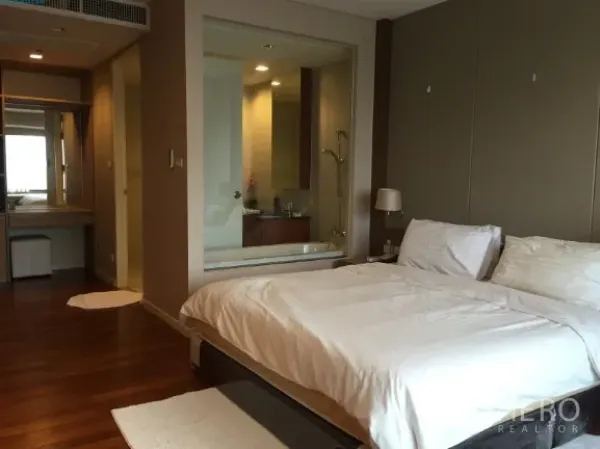 公寓 出租或出售在Sathon, Bangkok - 主卧配白色床品与木地板，玻璃墙浴室设浸泡浴缸与淋浴间。