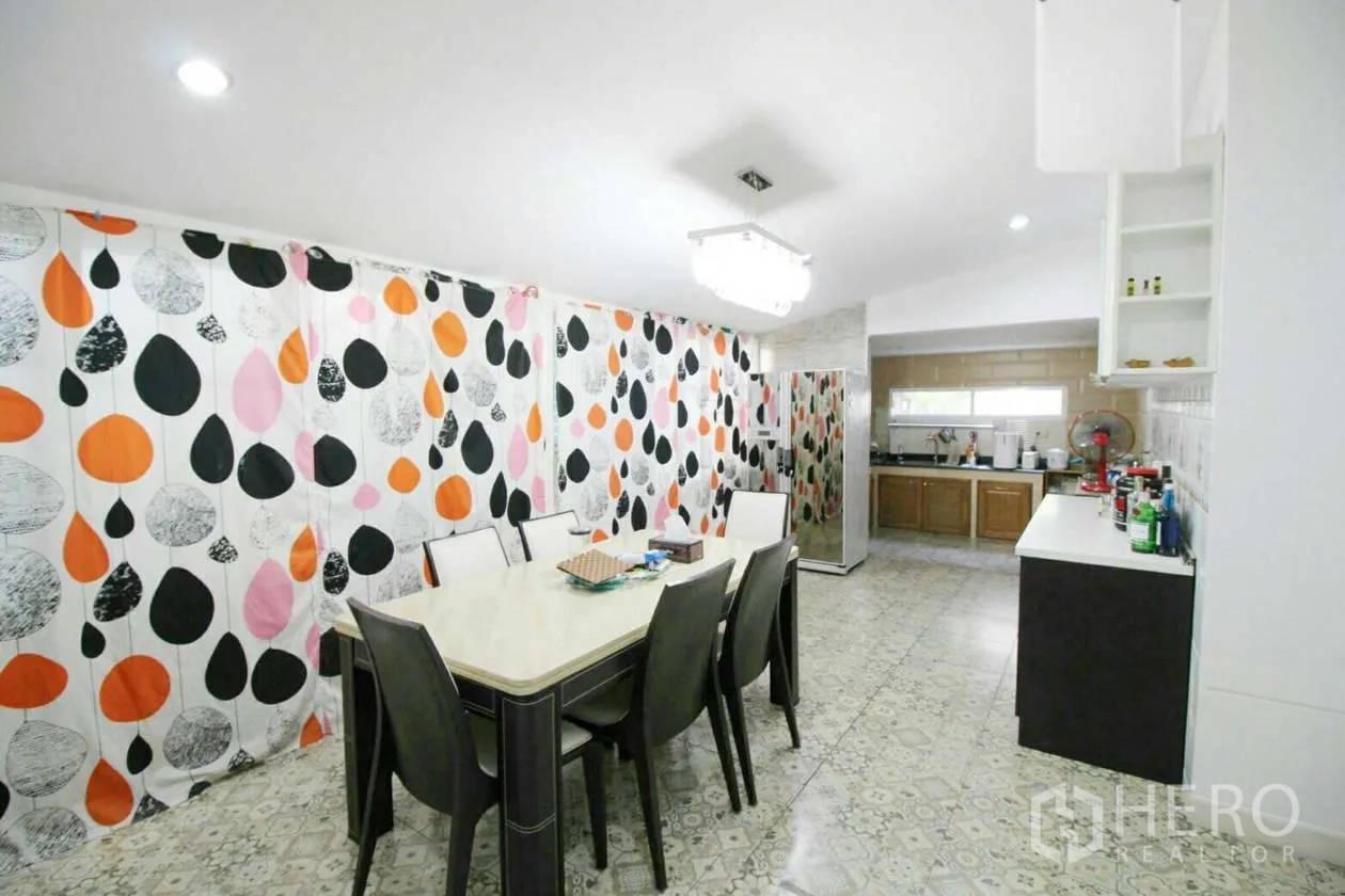 Detached House for rent or sale in Lat Krabang, Bangkok - Bright dining room with long table, colorful leaf-print curtains and adjoining kitchen.