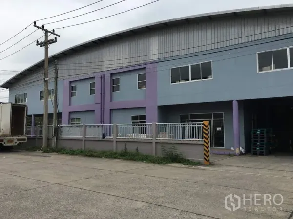 Factory for rent in Pathum Thani - Two-storey factory exterior with curved metal roof, blue and purple facade, fenced frontage and loading bay.