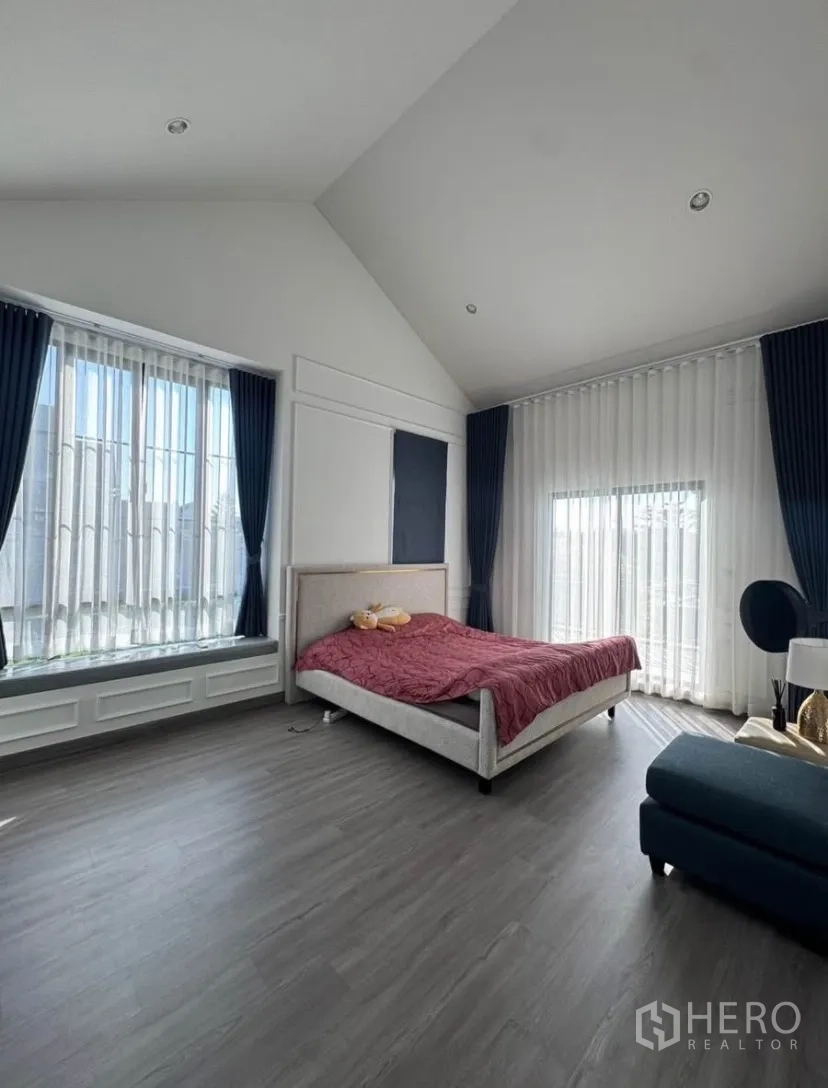 Detached House for rent or sale in Lat Krabang, Bangkok - Spacious bedroom with vaulted ceiling, floor-to-ceiling windows, sheer curtains, and a bed on grey wood floors.