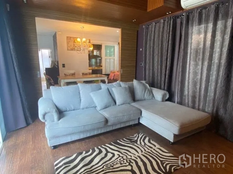บ้าน ให้เช่าหรือขายในPrawet, Bangkok - มุมนั่งเล่นอบอุ่น โซฟา L-shape พรมลายม้าลาย ผ้าม่านทึบ เห็นเชื่อมสู่ห้องกินข้าว