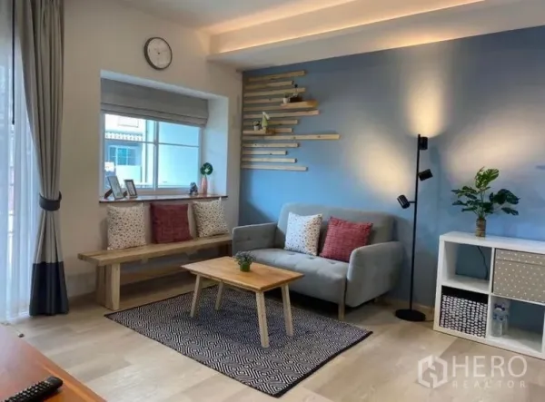 ทาวน์เฮาส์ ให้เช่าในBang Phli, Samut Prakan - ห้องนั่งเล่นสว่างของทาวน์เฮ้าส์บางพลี ผนังสีฟ้า ม้านั่งข้างหน้าต่าง โซฟาสีเทา และโต๊ะไม้เล็ก