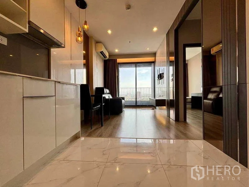 Condominium for rent or sale in Bang Na, Bangkok - Kitchen perspective toward the living room and balcony with marble-look floor tiles and warm lighting.
