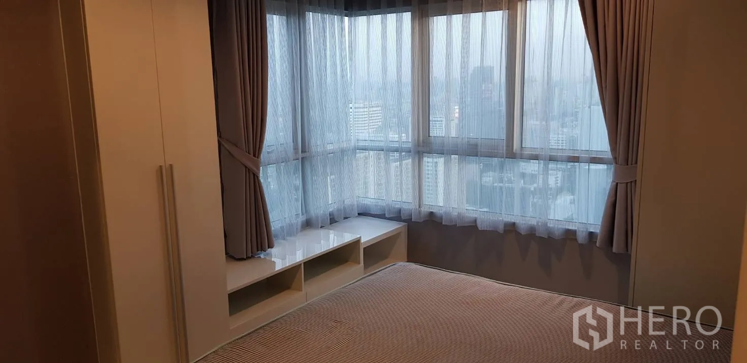 Condominium for rent in Huai Khwang, Bangkok - Master bedroom with corner windows, sheer curtains and wide city skyline views.