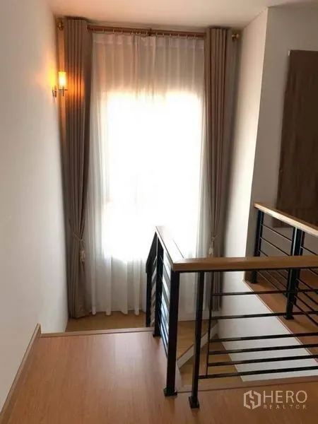 Detached House for sale in Suan Luang, Bangkok - Staircase landing with wooden handrail, black balusters and sheer curtained window.