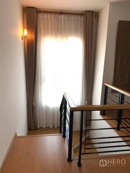 Detached House for sale in Suan Luang, Bangkok - Staircase landing with wooden handrail, black balusters and sheer curtained window.