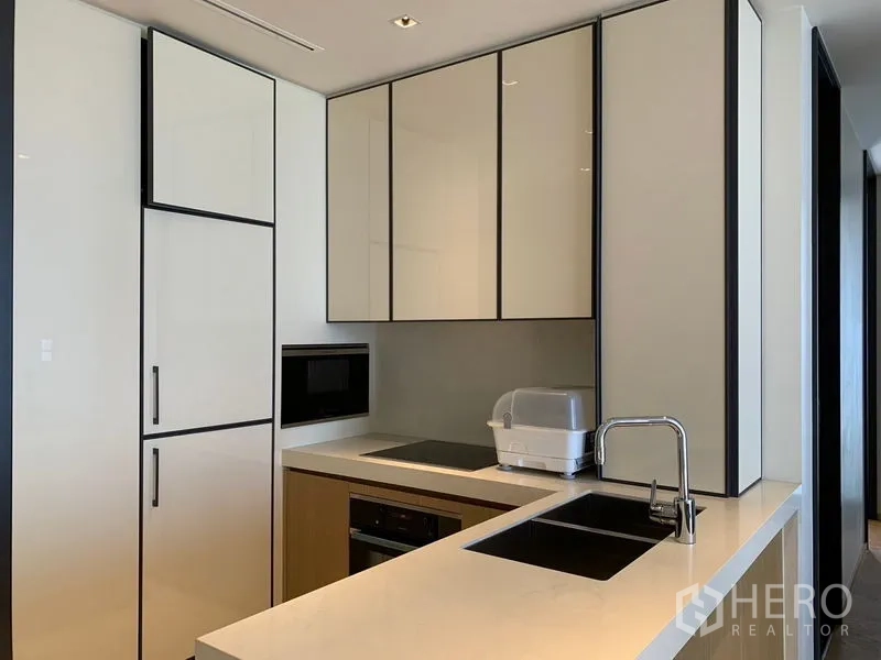 Condominium for rent in Khlong Toei, Bangkok - Modern fitted kitchen with white cabinets, built-in oven, cooktop, and sink on a long countertop.
