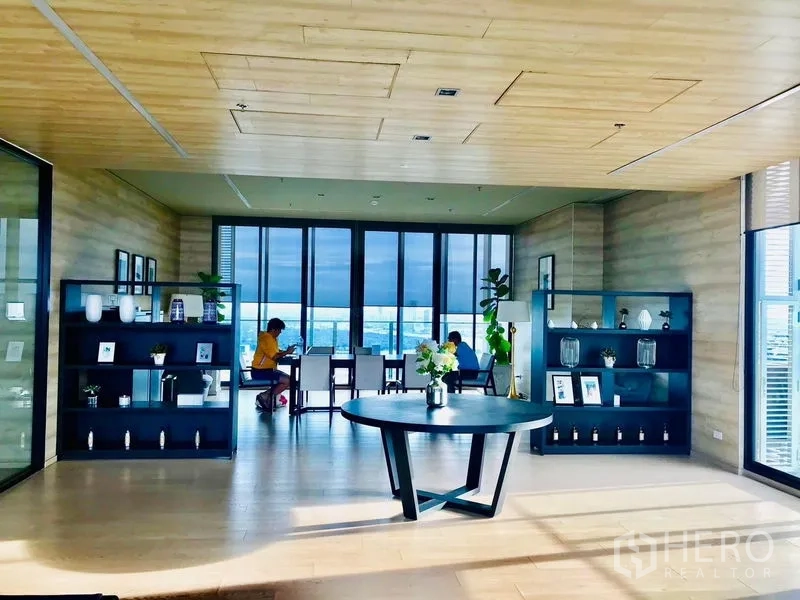 Condominium for rent in Khlong Toei, Bangkok - Residents’ lounge with wooden ceiling, black shelving and wide city-view windows beside a round table.