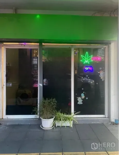 Shophouse for rent or sale in Khlong Toei, Bangkok - Street-level shopfront with sliding glass doors, green awning, neon leaf 'open' sign and potted plants.