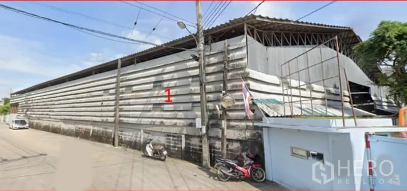 คลังสินค้า ให้เช่าในBang Khun Thian, Bangkok - มุมมองด้านหน้าตามถนนของโกดังผนังเมทัลชีทยาว พร้อมประตูทางเข้าและชายคากว้าง