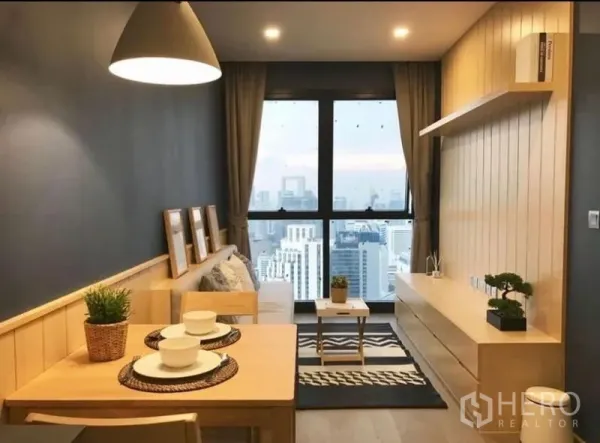 Condominium for rent in Watthana, Bangkok - Open-plan living and dining area with light wood built-ins, pendant lamp, and a large window showcasing a Bangkok skyline view.