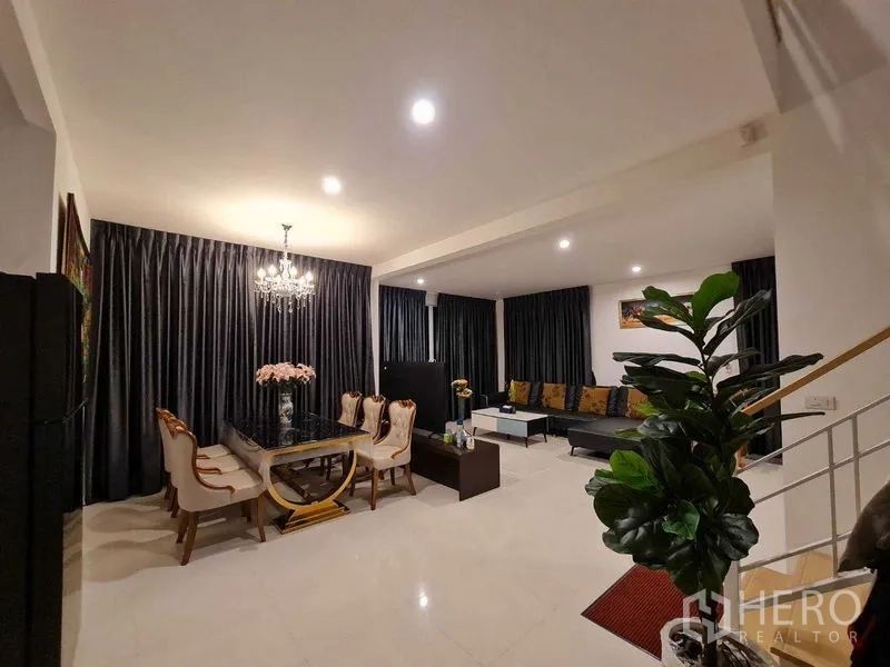 Detached House for rent or sale in Pathum Thani - Open-plan living and dining area with chandelier, marble table, leather seating, glossy tile floor and full-length curtains in a Thanyaburi detached house.