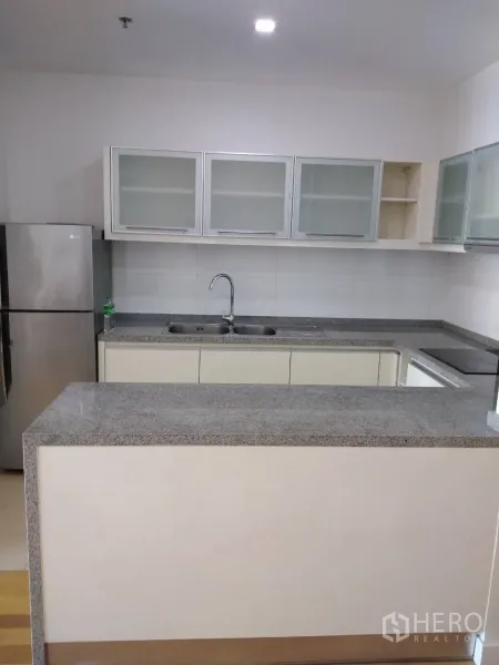 Condominium for rent in Khlong Toei, Bangkok - L-shaped kitchen with granite counters, double sink, glass-front cabinets, and stainless refrigerator.