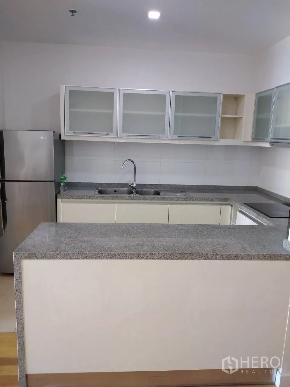 Condominium for rent in Khlong Toei, Bangkok - L-shaped kitchen with granite counters, double sink, glass-front cabinets, and stainless refrigerator.
