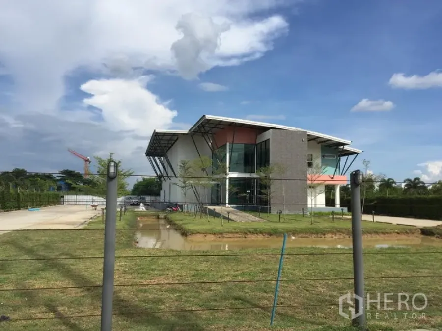 工厂 出租在Lam Luk Ka, Pathum Thani - Lam Luk Ka现代化工厂外立面，玻璃办公区、草坪、宽阔车道与水景，蓝天白云下。