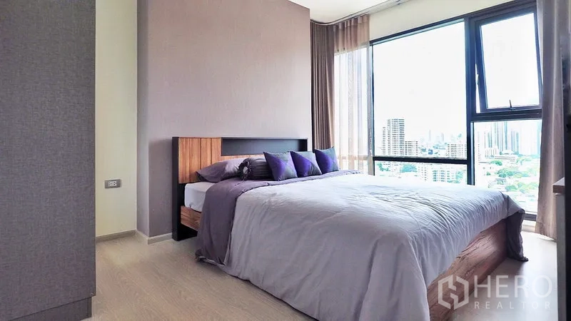 Condominium for rent in Khlong Toei, Bangkok - Corner master bedroom with large windows, city view and soft gray bedding.