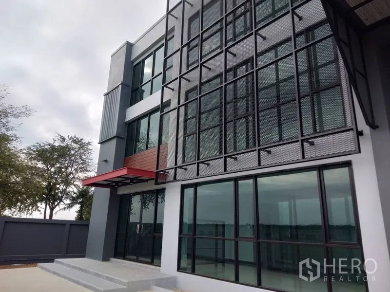 Factory for rent or sale in Lam Luk Ka, Pathum Thani - Close view of the facade showing floor-to-ceiling windows, metal sunshade grid and red canopy.