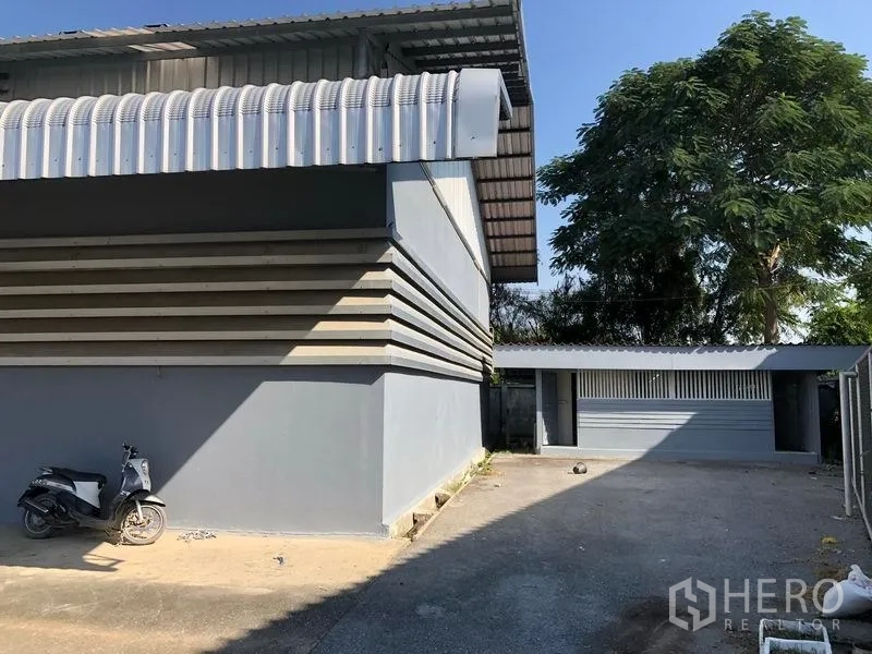 Factory for rent in Bang Bua Thong, Nonthaburi - Factory corner with side lane, mature tree, and a small auxiliary building.