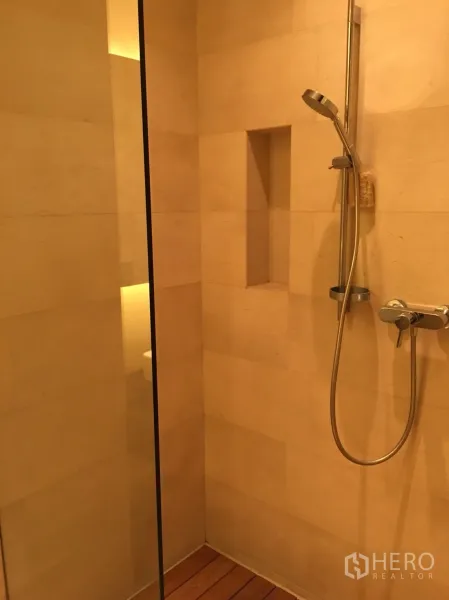 Condominium for rent or sale in Bang Rak, Bangkok - Walk-in shower with handheld showerhead, glass partition, built-in niche and warm stone tiles.