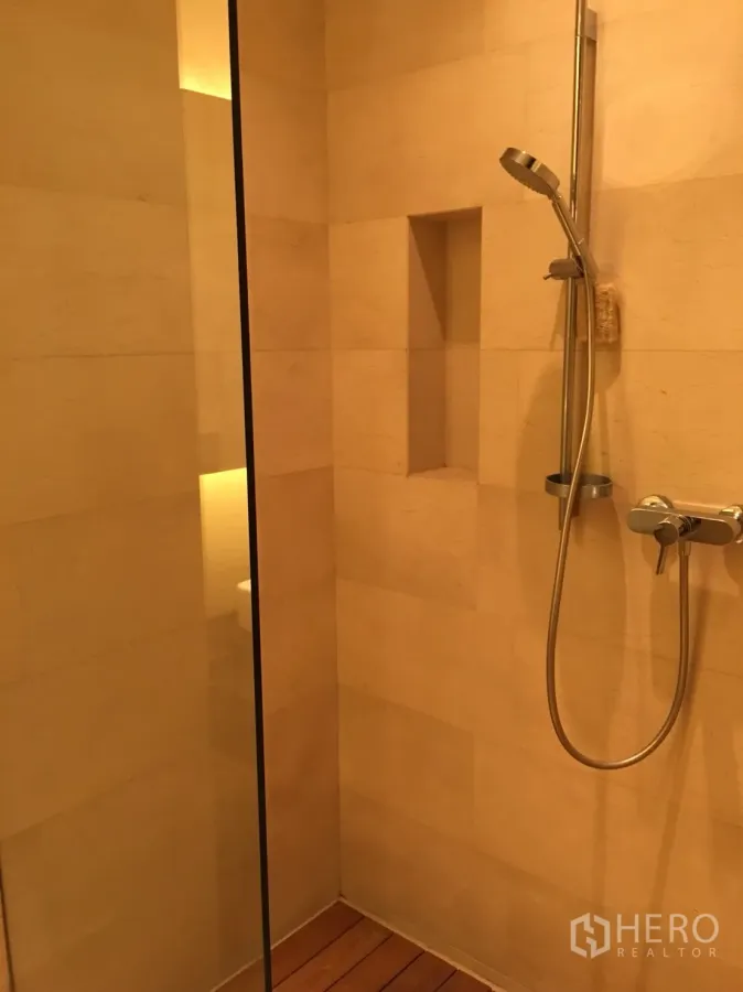 Condominium for rent or sale in Bang Rak, Bangkok - Walk-in shower with handheld showerhead, glass partition, built-in niche and warm stone tiles.