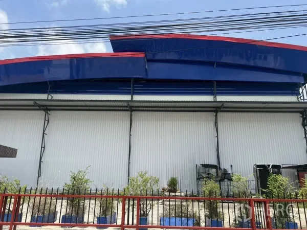 คลังสินค้า ให้เช่าในLat Krabang, Bangkok - ด้านหน้าคลังสินค้าในย่านลาดกระบัง หลังคาสีน้ำเงินตัดแดง ผนังเมทัลชีทสีขาว พร้อมรั้วและต้นไม้หน้าบ้าน.